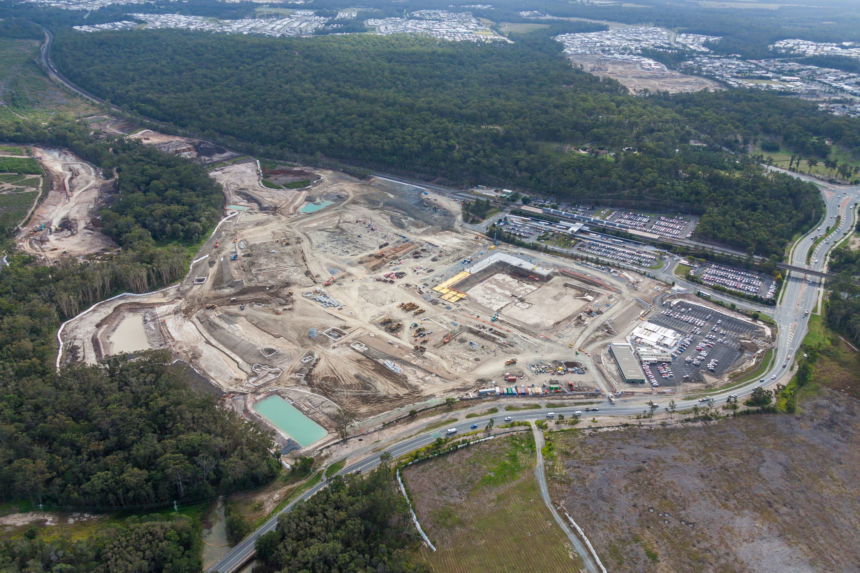 Aerial view of a large civil earthworks site with sediment basins and plant, beside a railway station car park and bushland