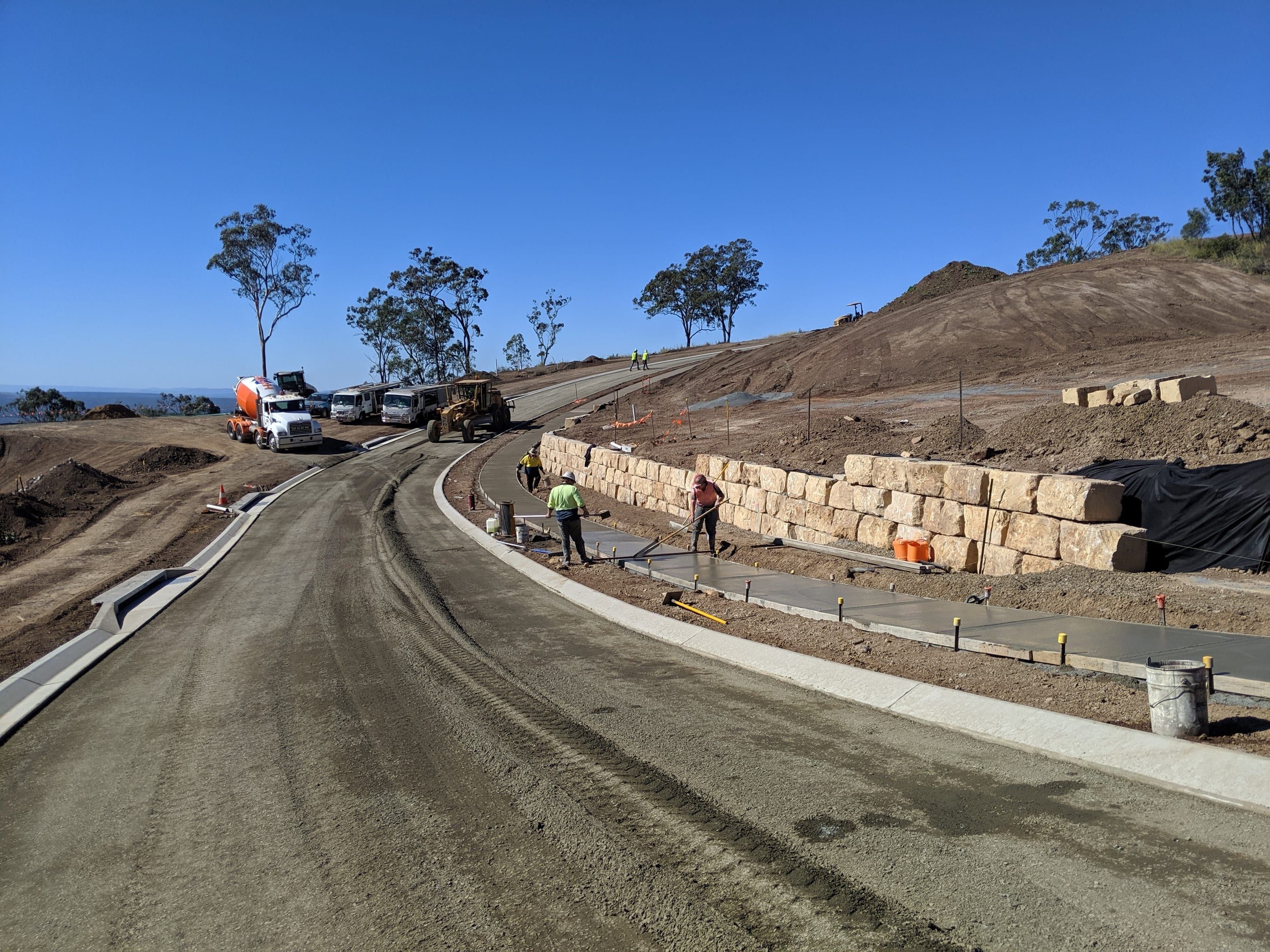 Crew finishing a concrete footpath beside a sandstone block retaining wall on a new subdivision road